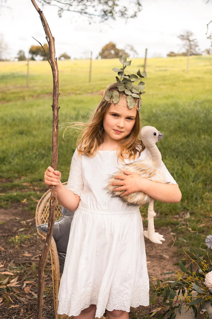 A young girl in a white dress stands outdoors on a grassy field, holding a long wooden stick with one hand and cradling Eddie The Emu Stuffed Animal, suitable for ages 3+, in the other. With a crown made of leaves on her head and wearing a serious expression, she poses against the backdrop of trees and a fence.