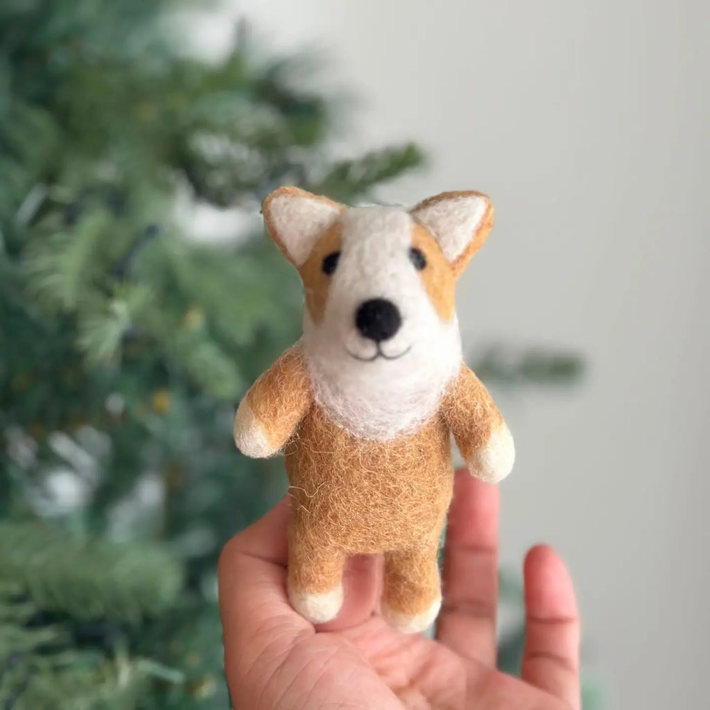 A charming Corgi Dog Finger Puppet, handcrafted from 100% wool, with brown and white fur, delicately perched on a finger. The backdrop showcases a softly lit, out-of-focus green Christmas tree.