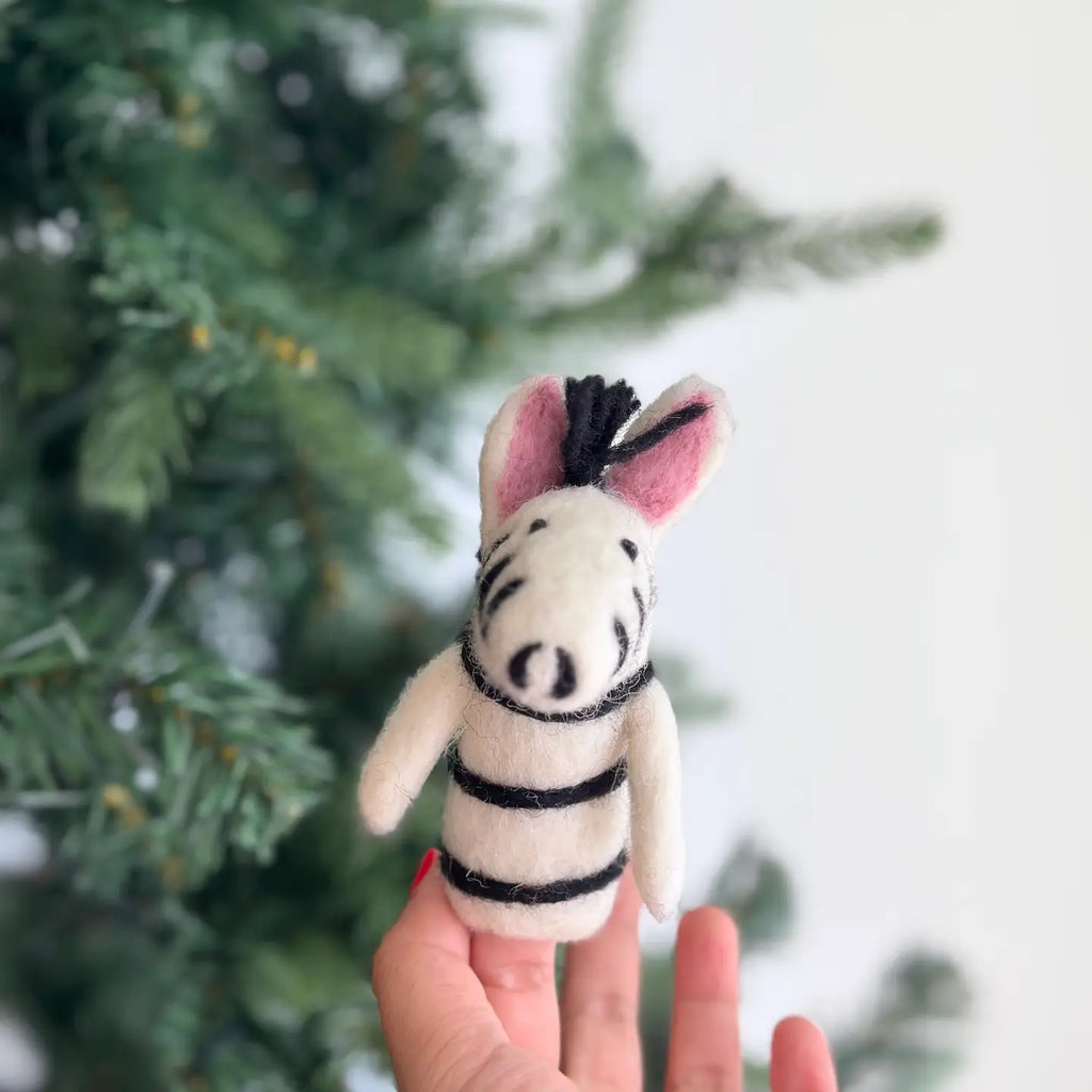 A person holds up a small Zebra Finger Puppet with a pink nose and ears, black stripes, and a mane. Wild animal lovers will adore this charming piece. In the background, there is a blurred image of a Christmas tree.