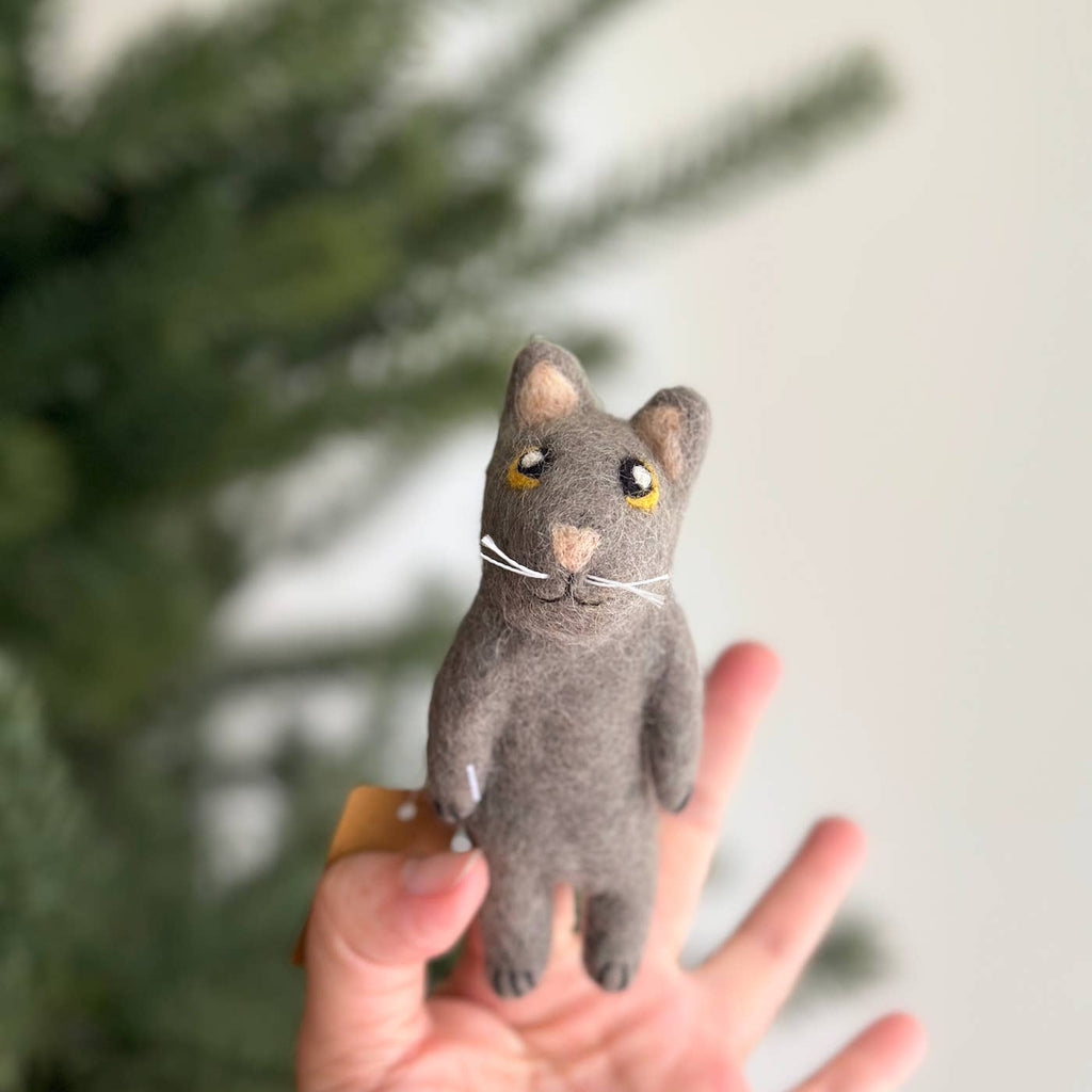 A hand holds the Felt Gray Cat Finger Puppet, a small, gray creation with yellow eyes and white whiskers. The backdrop is filled with softly blurred green branches, likely of a tree.