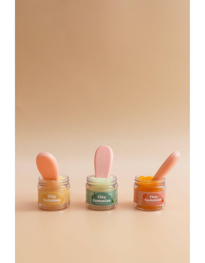 Three small jars of Tiny Tummies - Banana Jelly food by Tiny Harlow in different colors are arranged in a row against a beige background. Each jar has a magic spoon placed inside, resembling a setup for when a doll enjoys its meal.