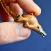 A hand holding a delightful Platypus Necklace, showcasing a charming 24k gold plated steel chain with a small ceramic platypus pendant against a blue background. The handcrafted porcelain pendant features an orange and beige platypus with detailed flippers, bill, and nose.