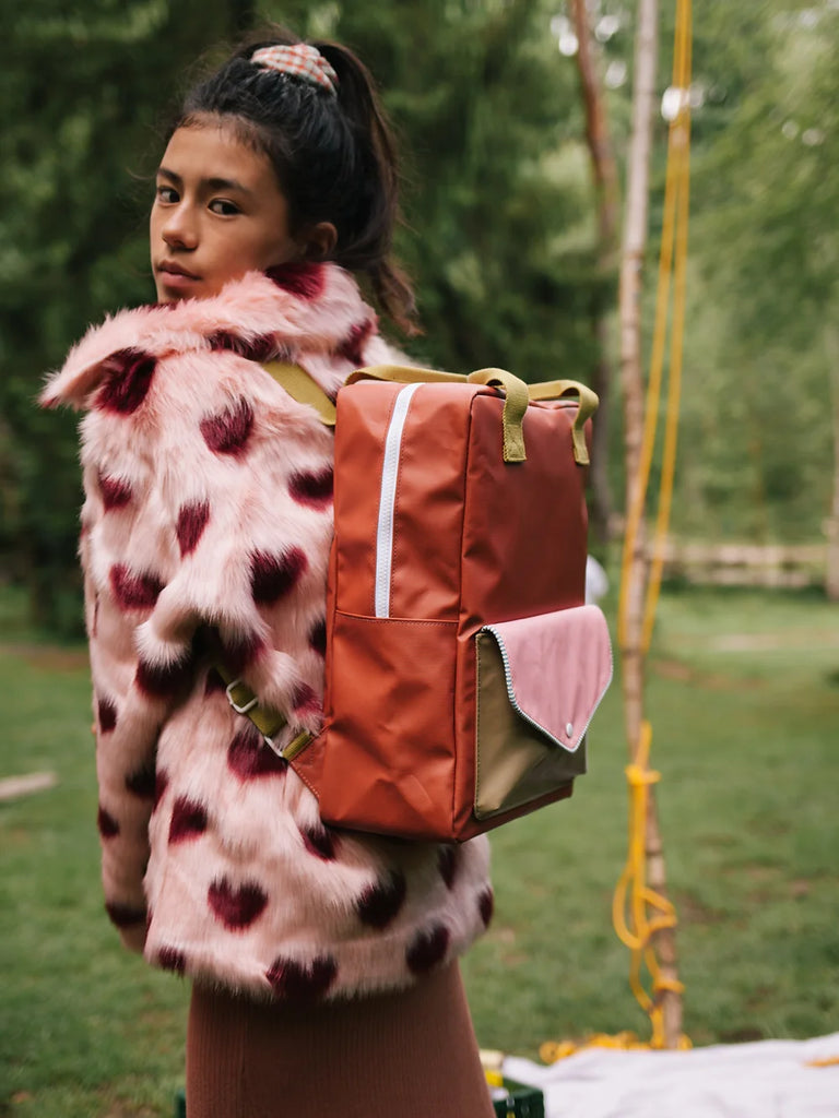 A young girl in a colorful faux fur coat with a Sticky Lemon Backpack Large from the Envelope Collection in Lighthouse Red made of waterproof nylon looks over her shoulder in a lush green park setting.