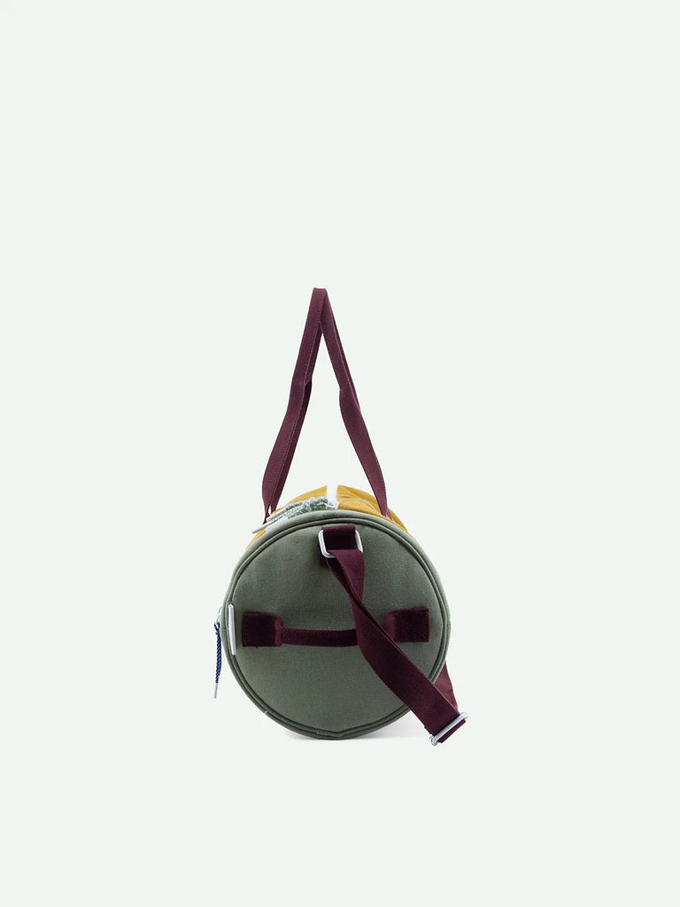 A circular Sticky Lemon Duffle Bag in Khaki Green, with burgundy straps, positioned upright against a white background. The bag features a side zipper and a visible top handle.