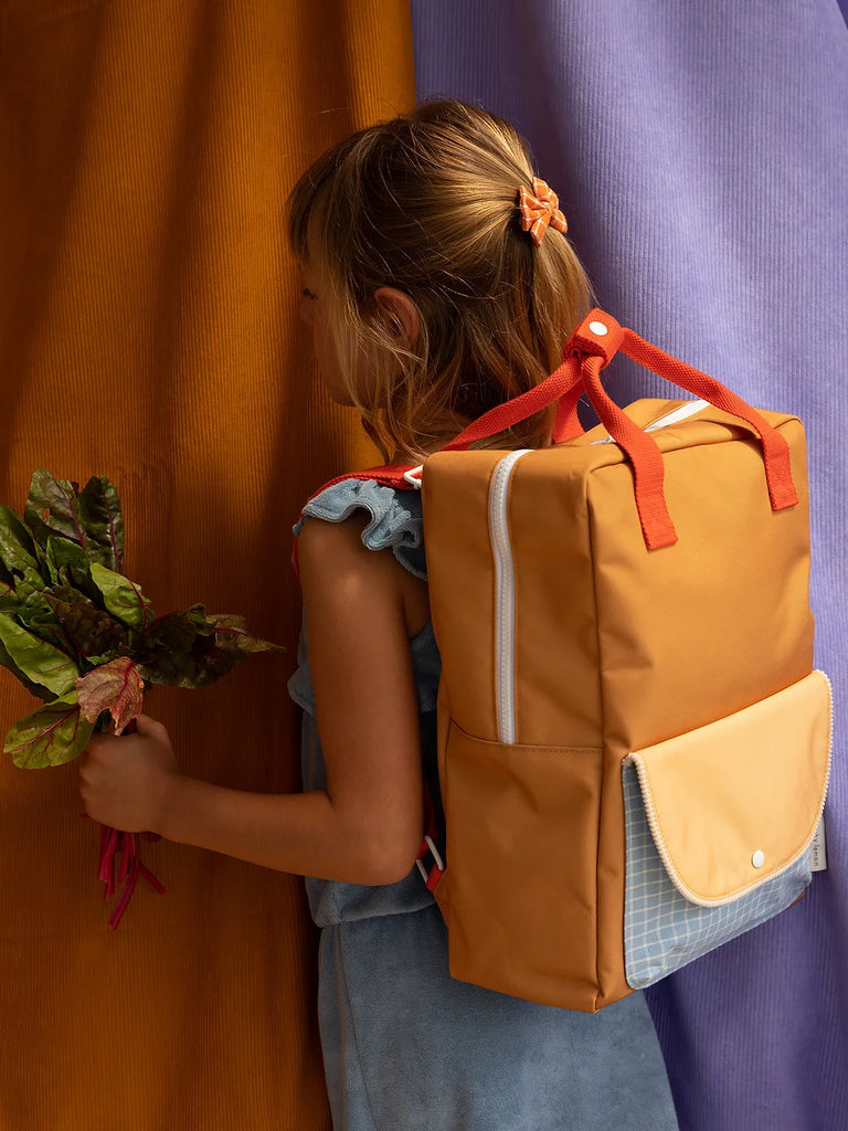 A young girl with a Sticky Lemon Backpack Large holding a bouquet of flowers, standing against a colorful fabric background.