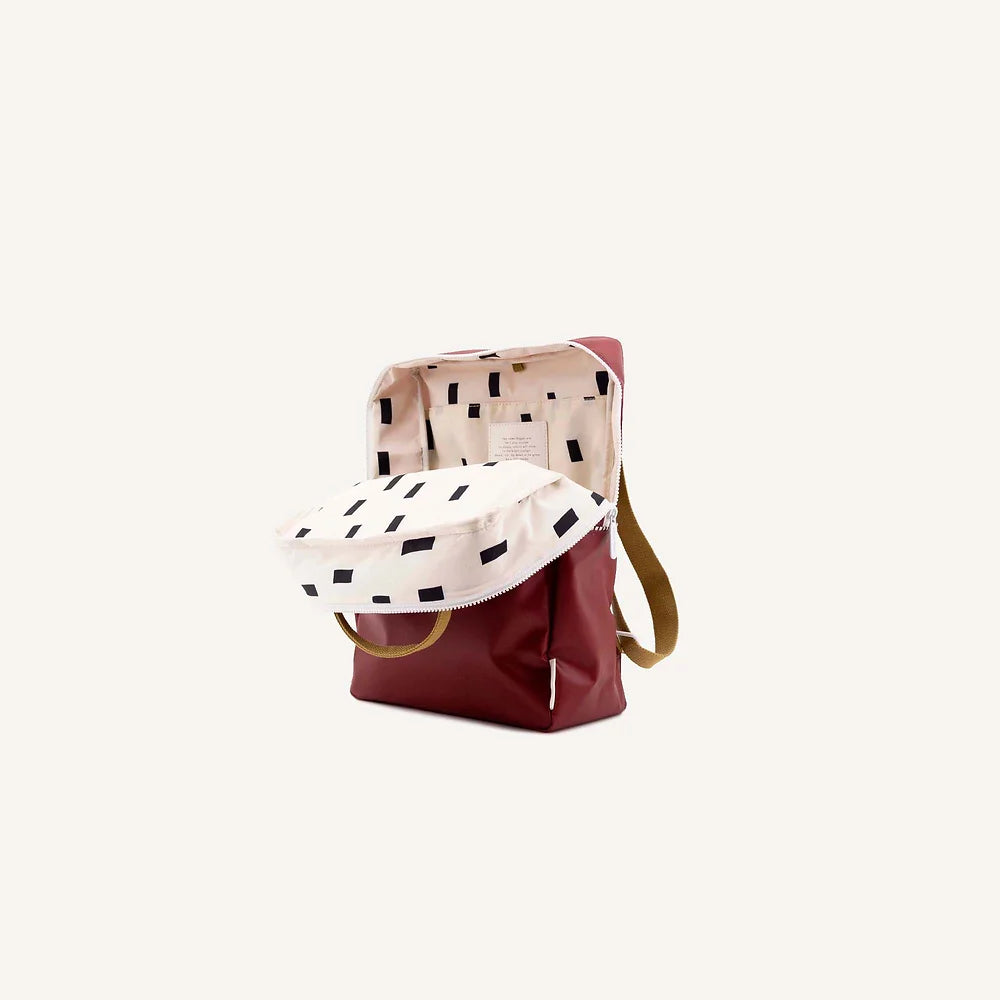 A stylish Sticky Lemon Backpack | Uni | Journey Red made of waterproof nylon, with a beige upper half decorated with black dashes and a solid maroon lower half, featuring a beige handle and straps, set against a plain white background.