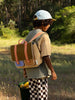 A young boy wearing a cap, backpack, and colorful outfit stands in a sunny field with a Corduroy Pear Jam shoulder bag of fruit, gazing towards the trees.