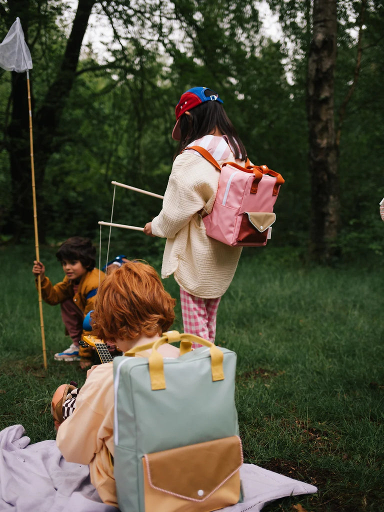 Three children in a forest setting, two sitting on a blanket and one standing, all holding sticks. The standing child, wearing a Sticky Lemon Backpack Large from the Envelope Collection in Map Green made from recycled PET bottles, faces away. They seem to be