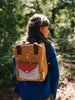 A young girl stands in a forest wearing a blue sweater and carrying a Sticky Lemon Backpack Small Special Edition | Adventure Collection | Khaki Green with playful patterns, including acorns and small creatures. The sunlight filters through the trees, creating a serene natural backdrop.