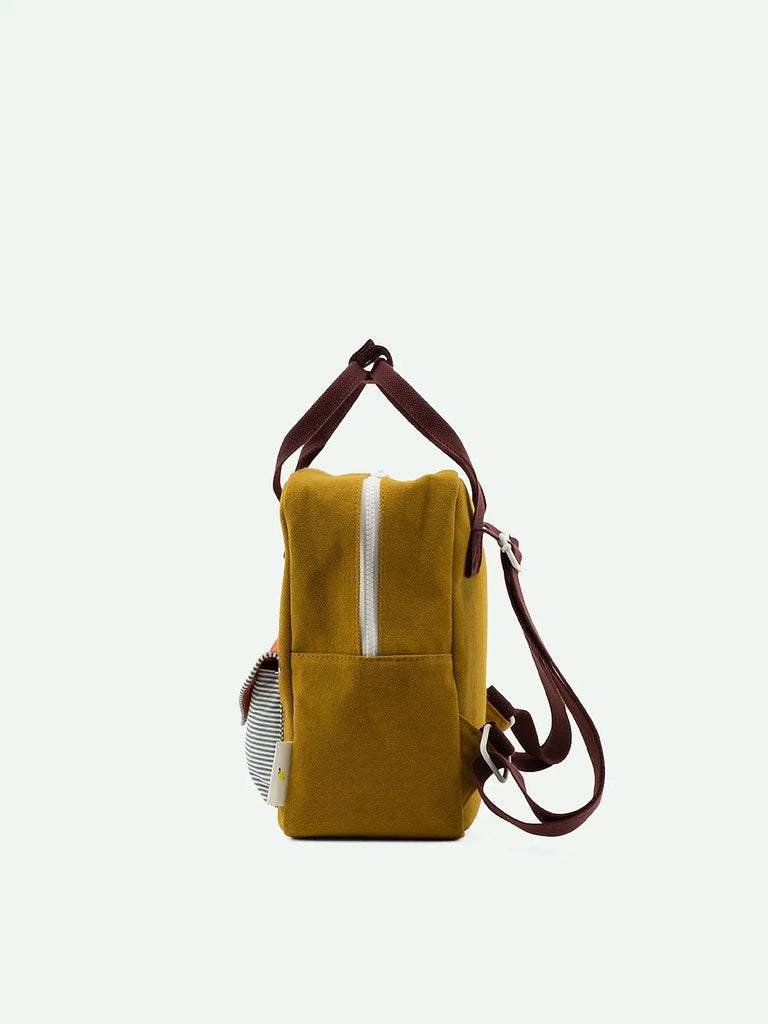 Side view of a mustard yellow canvas bag with a white zipper and brown straps. The Sticky Lemon Backpack Small Special Edition | Adventure Collection | Khaki Green has a small side pocket with a striped item peeking out and features a sturdy shoulder handle. The background is plain light grey.