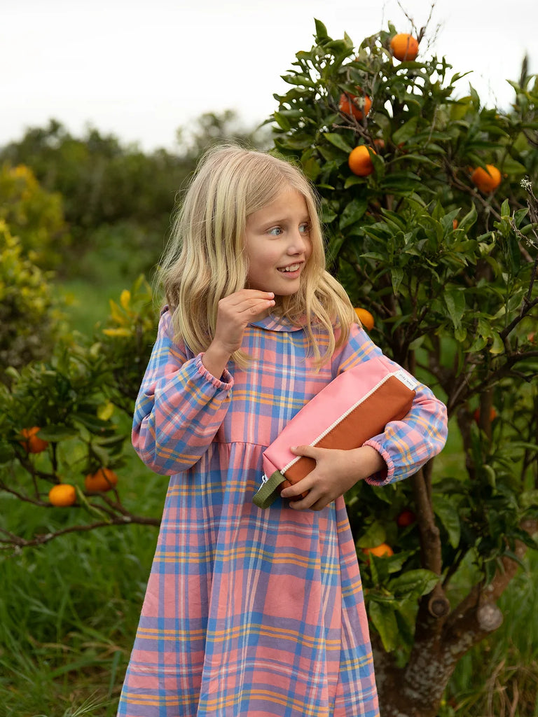 A young girl with long blonde hair, dressed in a pink and blue plaid dress made from recycled PET bottles, stands in front of an orange tree. Clutching a Sticky Lemon Pencil Case in Flower Pink and Willow Brown to her chest, she looks to her left with a slight smile. The background showcases more trees filled with ripe oranges and lush greenery.