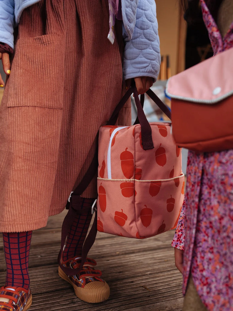 Two children standing close together, with a focus on their lower bodies. One child holds a Sticky Lemon Backpack Small | Envelope Collection | Special Edition Acorn, wearing orange textured overalls and patterned socks with brown shoes.