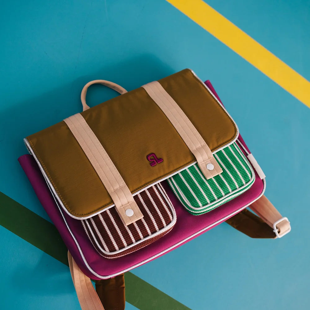 The Sticky Lemon School Bag | Better Together | Gymnastic Pink, designed with a colorful pattern and made from waterproof nylon, is placed on a diagonally-striped blue surface. It features a brown top flap with tan straps, a pink body, and two front pockets adorned with brown and green vertical stripes. A small handle at the top along with padded shoulder straps complete its look.