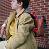 A young person with short hair sits against a brick wall. They wear a light brown coat over a yellow and white striped sweater and carry the Sticky Lemon School Bag | Better Together | Leather Ball made from recycled bottles, featuring red and blue straps, over their shoulder. The person gazes to the side with a contemplative expression.