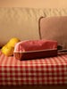 A pink and brown rectangular pouch, specifically the Sticky Lemon Pencil Case in Flower Pink + Willow Brown, lies on a red-and-white checkered surface. Next to it, two lemons rest on the same surface. In the background, a beige sofa is adorned with a brown cushion. The pencil case, made from recycled PET bottles, adds an eco-friendly touch to this cozy scene.