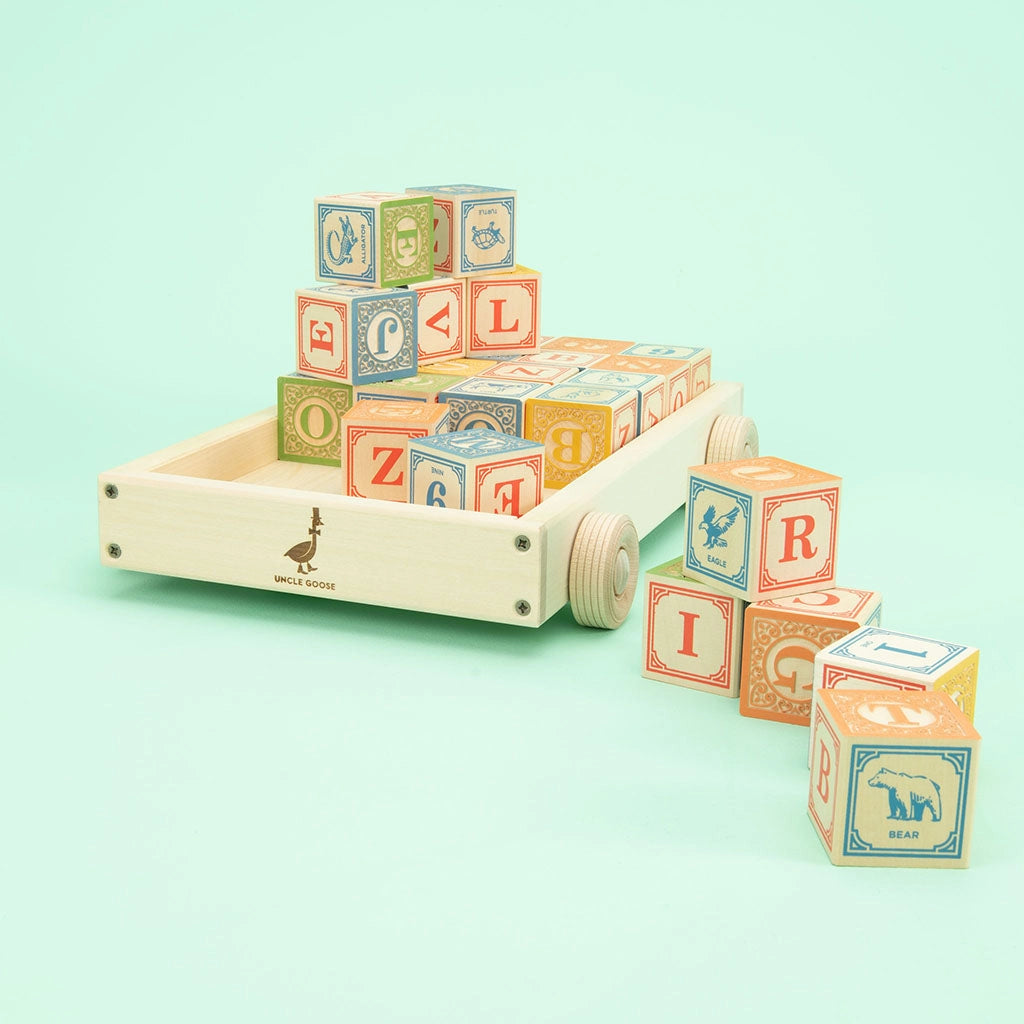 Uncle Goose Classic ABC Blocks with Wagon with letters, numbers, and animal illustrations, some stacked in a handmade basswood wagon and others scattered around. The blocks are colorful, and the wagon has four wheels. The background is a light mint green color.