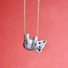 Introducing our Koala Necklace: This charming pendant necklace showcases a small, hand-painted ceramic koala with intricate white and gray fur details. The playful koala hangs upside down from a gold plated chain, set against a solid coral pink background.