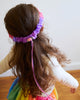 A young child with long brown hair is seen from behind, wearing a Sarah's Silk Blossom Garland Headband. The child, perhaps engaged in dress-up play, is dressed in a multicolored outfit and stands on a wooden floor against a plain white wall adorned with delicate garlands.