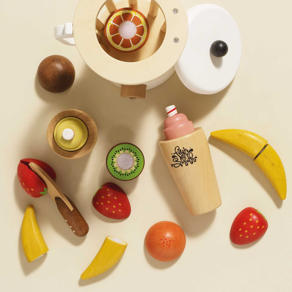 The Fruit & Smoothie Blender Set features a selection of sliceable wooden toy fruits, such as bananas, strawberries, and a kiwi slice, arranged around a vintage-style wooden juicer. Included is also a charming wooden bottle designed to resemble a milkshake or smoothie, all set against a light cream background in this delightful toy kitchen scene.