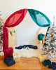 A festive setup includes a children's play tent adorned with Sarah's Silk Giant Evergreen Playsilk in vibrant red and green, surrounded by small, flocked Christmas trees. Wrapped gifts in winter colorways of red, green, navy, and cream are placed on a fluffy white rug, enhancing the holiday ambiance.