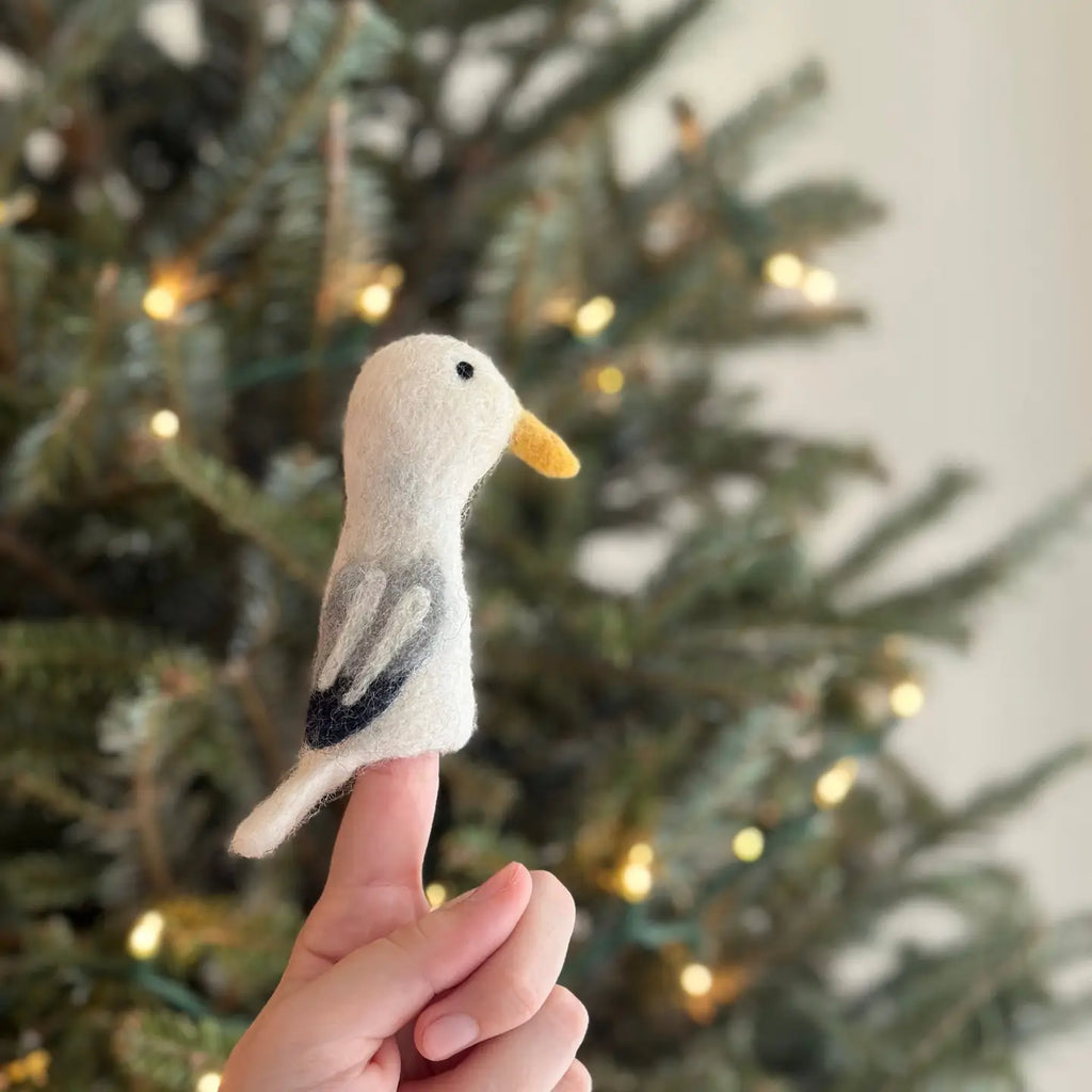 The Seagull Finger Puppet, a small handmade creation with white feathers and a yellow beak, is perched delicately on an index finger. In the background, an out-of-focus Christmas tree decorated with warm, glowing lights provides a festive ambiance.