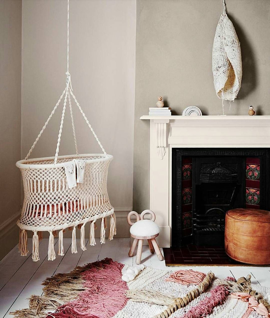 A cozy room corner featuring a hanging macramé cradle next to a fireplace, a small wooden stool, a round orange ottoman, and a colorful textured faux sheep chair on a white floor.