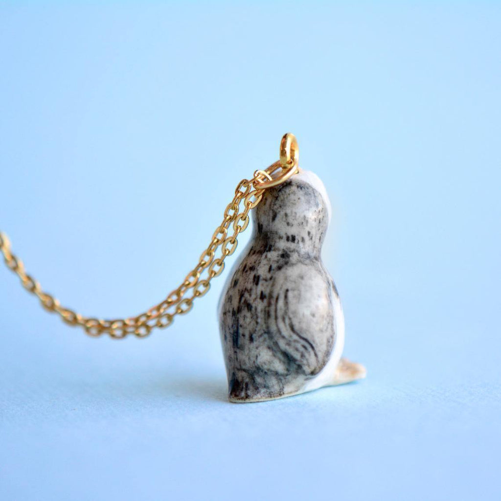 A Tiny Penguin Necklace, photographed against a blue background. The handmade necklace features the penguin facing away, showcasing its gray and white textured back.