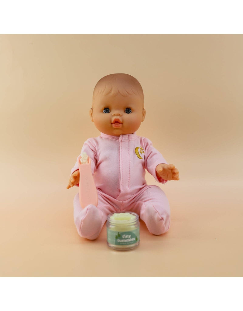 A baby doll dressed in a pink outfit sits against a peach-colored backdrop. It holds a small bottle, with a jar of "Tiny Tummies- Banana Jelly food" by Tiny Harlow placed in front, ready for the little one to enjoy.