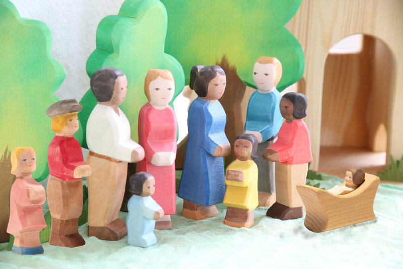 A vibrant assembly of wooden Ostheimer figurines, featuring adults and children, stand arranged in a semi-circle on a green surface. In the foreground, a baby figurine rests peacefully in a wooden cradle. Made from sustainable materials, these handcrafted toys include charming details such as the Ostheimer Son with Gray Hat and are complemented by trees and a wooden structure in the background.