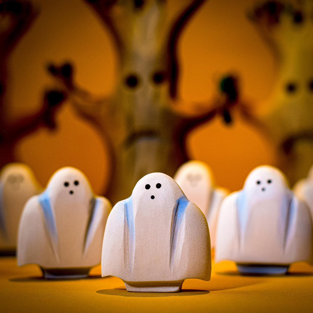 A group of small, Handmade Wooden Ghost figurines with two black holes for eyes and one for a mouth are arranged in front of a blurry background featuring a large tree with facial features. The scene, perfect for a spooky story, is illuminated with a warm, yellowish light.