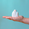 A small, white Handmade Wooden Ghost with brown eyes sits on the outstretched palm of a person's hand, ready to embark on a playtime adventure. The background is a solid, light blue color.