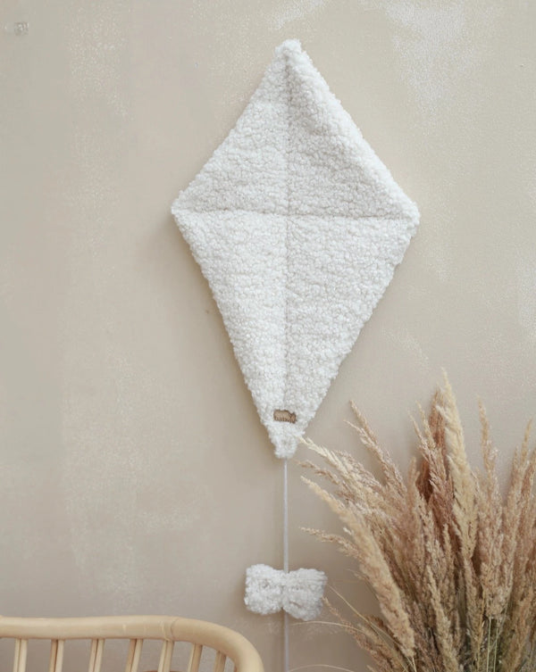 A Boucle Kite Wall Decor - Cream hangs on a beige wall above a wicker chair. The kite features a soft, fluffy texture with two small bows attached to its tail—ideal for children's room decoration. Beside the chair, there is a bundle of dried pampas grass in natural tones.