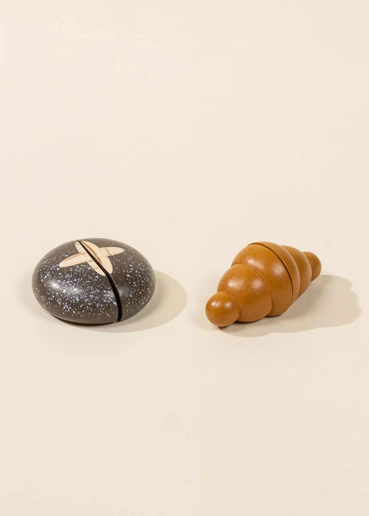 A piece from the Wooden Bakery Set, featuring a brown croissant-shaped item and a round, black, speckled object adorned with a wooden butterfly design on top, are displayed against a beige background, evoking elements of a children's game.