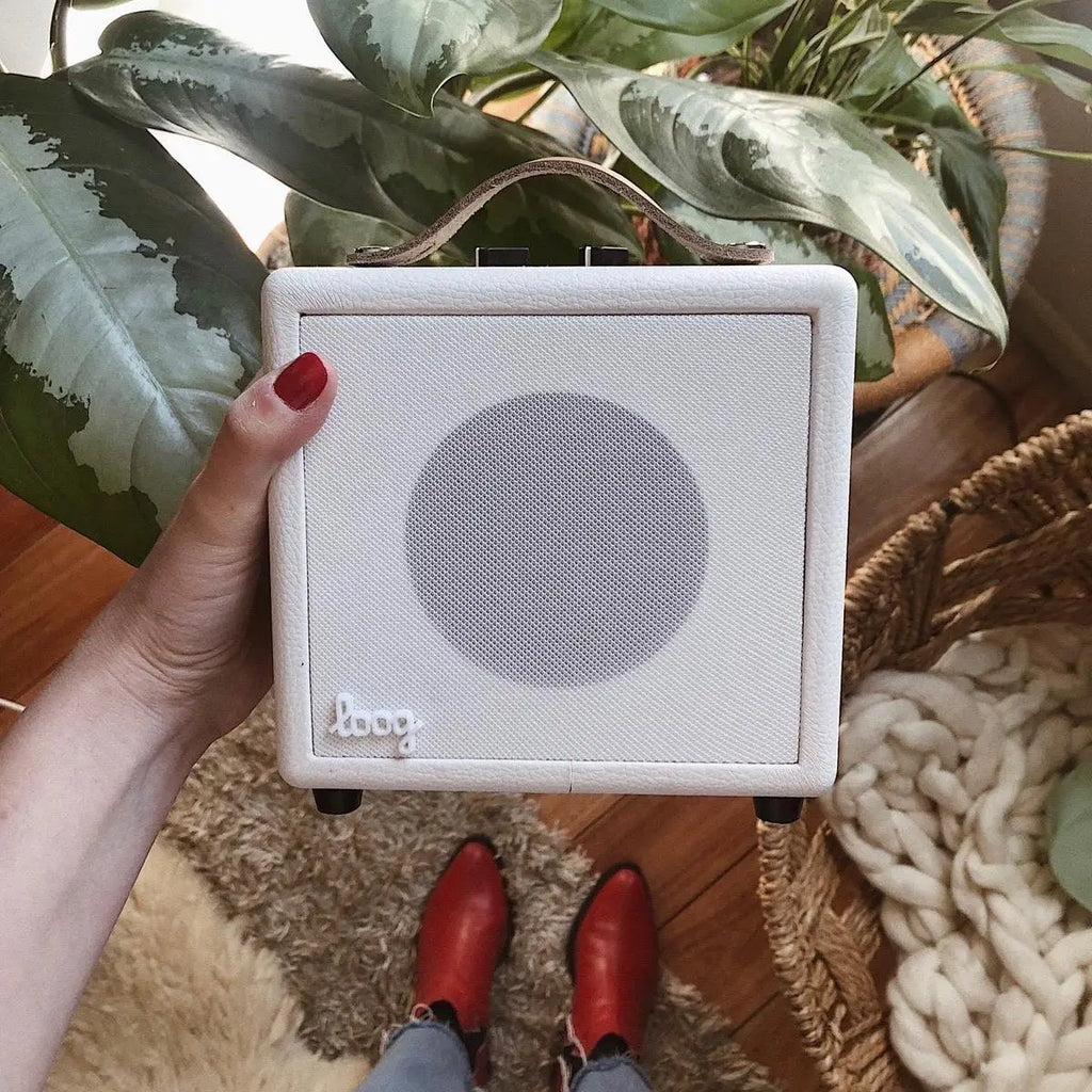 A person holds a small, white portable speaker branded "Mini Guitar Amp" in one hand against an indoor background with green plants and a white textured rug. They wear bright red boots.