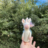 A hand holds the Unicorn Finger Puppet, a small, whimsical felted wool figurine with a unicorn horn and wings, set against a backdrop of lush green foliage and a blue sky. This needle-felted masterpiece is crafted from 100% wool.