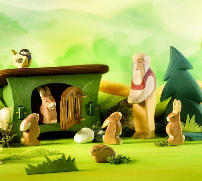 A whimsical scene of handcrafted wooden toys, perhaps by Ostheimer, depicts an Ostheimer Grandfather standing near a small house with a green roof, which has a rabbit inside. Surrounding him are several other rabbits, trees, and a bird perched on the house. The background is a soft, green landscape.