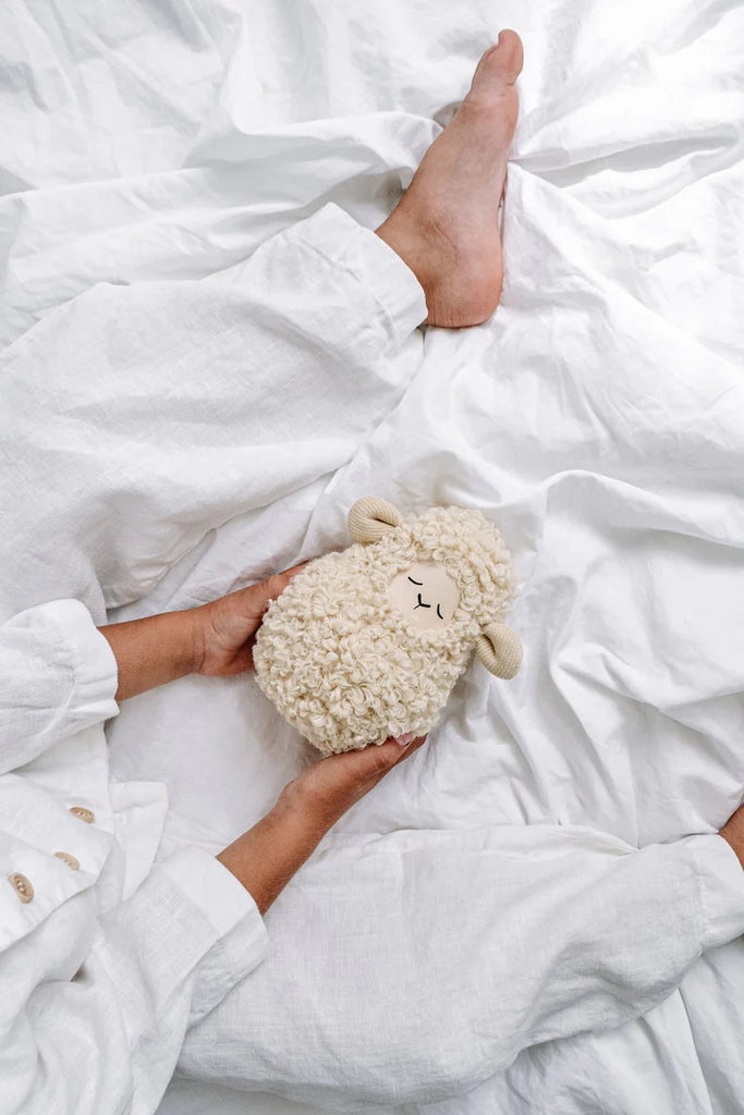 Person holding a Handmade Musical Roly Poly - Lamb toy while sitting on a bed with white sheets and visible bare feet.