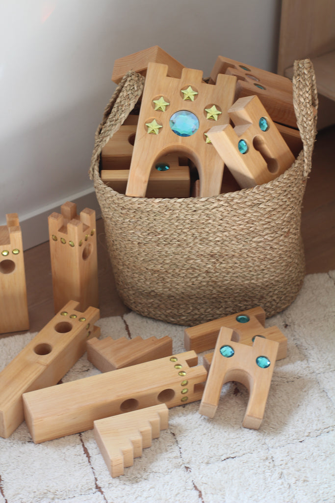 A woven basket filled with wooden building blocks, some decorated with blue gem-like and gold star stickers, sits by a white wall. Several blocks from the Bauspiel Large Fairytale Castle (16 pieces) set are arranged on the light-colored rug, inviting imaginative play.