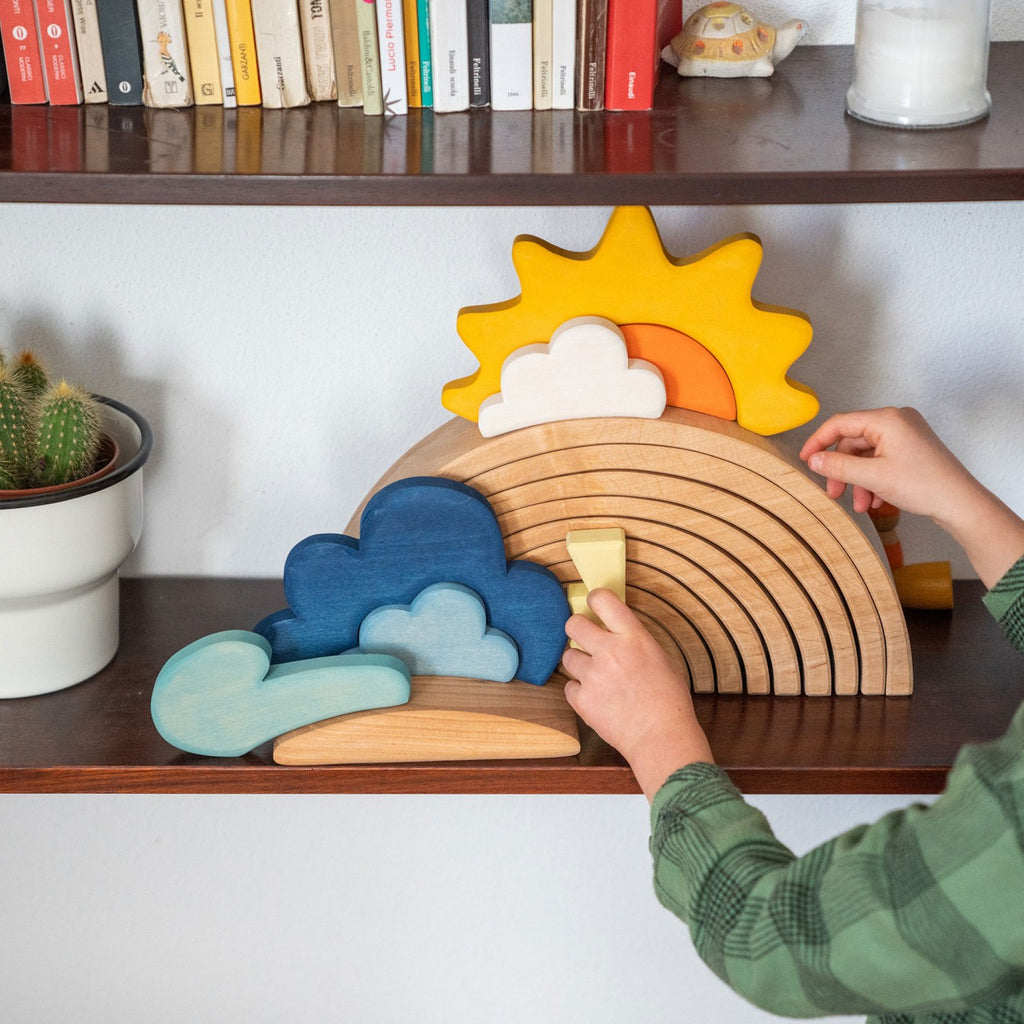 A child wearing a green plaid shirt assembles a Grimm's Weather Building Block Set featuring a sun, clouds, and rainbow arches. The puzzle, perfect for imagination development with children toys, is placed on a wooden shelf adorned with various books and a potted cactus.