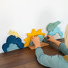 A child is playing with the Grimm's Weather Building Block Set on a dark wooden surface. The toys include shapes representing a cloud with a lightning bolt, the sun, and clouds. As the young builder arranges the blue cloud shapes on a small wooden base, they are weaving their own weather stories.