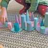 A child wearing a green shirt and red polka dot pants plays with the Pastel Semi Circle Set on a patterned rug, reminiscent of modern architecture. The shapes include cylinders, cones, and discs in pastel shades of blue, pink, and purple, arranged in a symmetrical pattern.