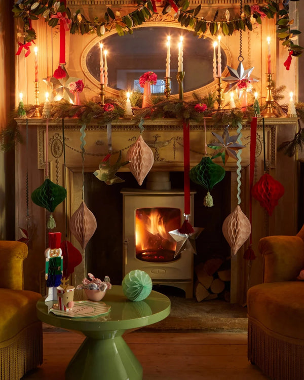 A cozy living room features a lit fireplace with a decorative mantle adorned with candles, stars, and colorful paper ornaments. Warm lighting enhances the festive backdrop. A small green table sits in front, boasting a nutcracker and elegant MeriMeri Large Hanging Decorations.