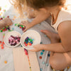 Two young children play with colorful mosaic tiles, arranging them on Grapat Mix & Match educational toys that include wooden sorting boxes. They are seated on a light fabric surface surrounded by scattered tiles and bowls filled with more pieces.