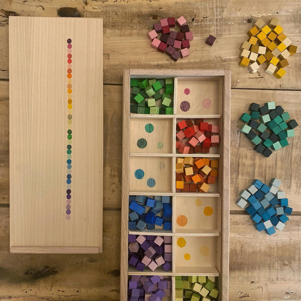 A Grapat Mix & Match sorting box with a column of colorful spots next to a tray containing compartments filled with multicolored wooden blocks, some scattered on a table.