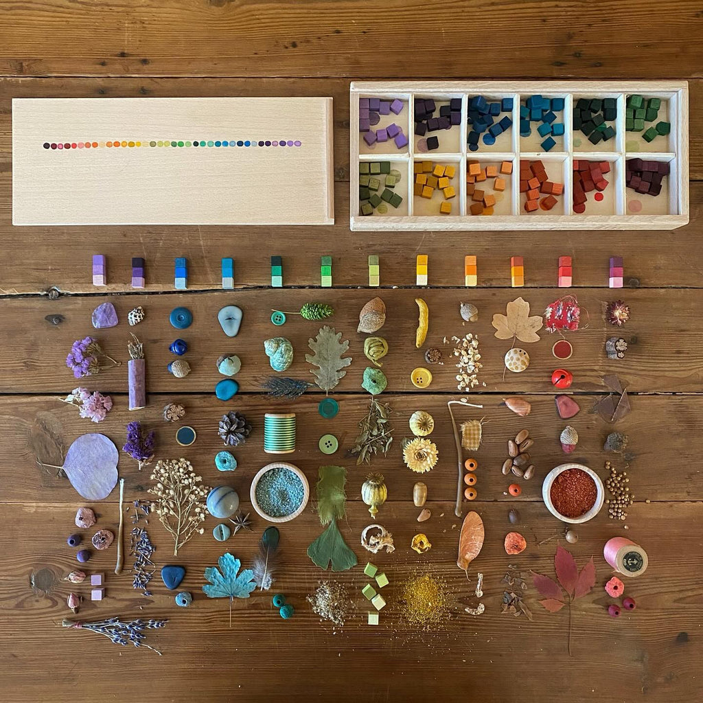 A wooden table displays an assortment of Grapat Mix & Match beads, buttons, natural elements, and colorful cubes in various shapes neatly arranged.