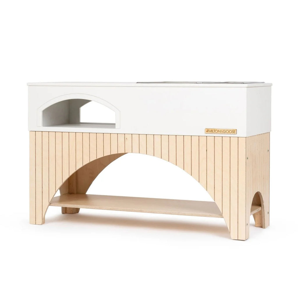 A modern wooden kids' kitchen step stool by Milton & Goose Mud Kitchen (Made in USA), with a white top and natural wood base. It features a cut-out step on the lower half and an adjustable platform on top, designed to help children reach higher surfaces safely. Perfect for both indoor use and alongside an outdoor playset.