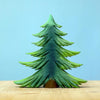 A vibrant green, three-dimensional model of a Bumbu Large Wooden Fir Tree stands on a light wood surface. Handmade in Romania, the tree boasts layered, angular branches and is set against a soft blue background.