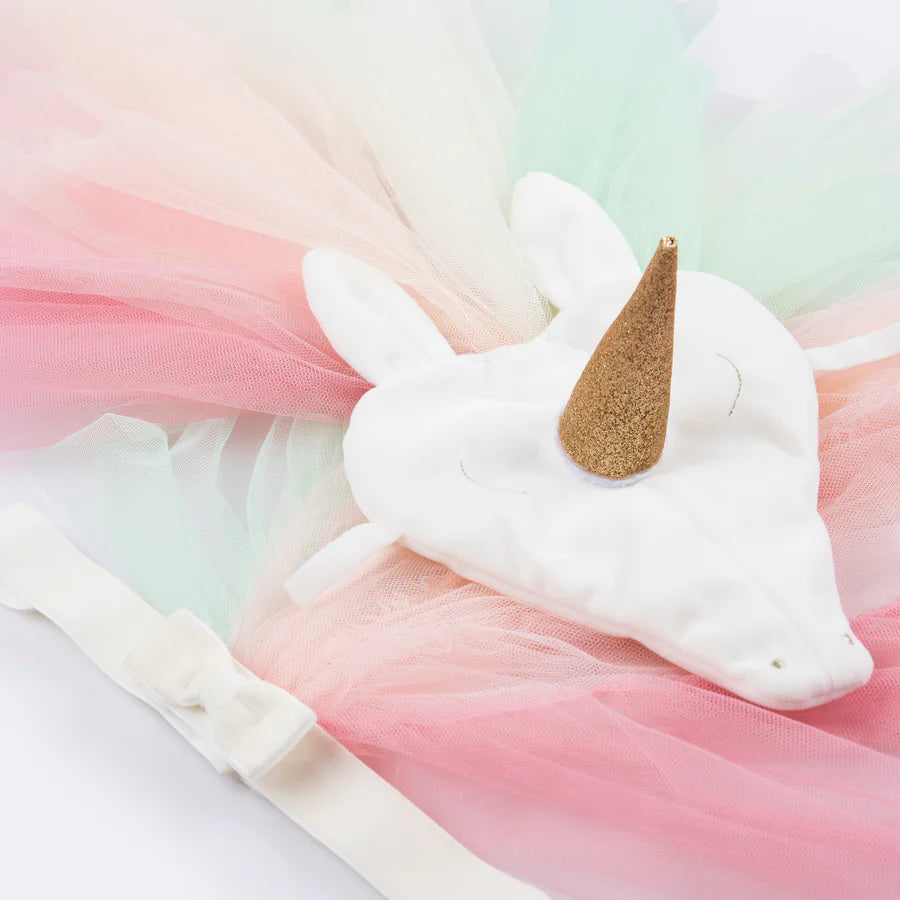 The Meri Meri Rainbow Unicorn Costume - Final Sale features a white unicorn mask adorned with a golden horn and white ears, surrounded by layers of pastel pink, green, and blue tulle fabric. This captivating dress-up set also includes a flowing rainbow tulle tail and a matching velvet unicorn headpiece. A white ribbon is prominently visible in the foreground.