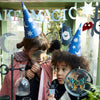 Two children in Meri Meri Wizard Party Hats, one holding a magnifying glass to a butterfly, the other peering through a magnifying glass at a small toy. They are surrounded by Halloween party decorations and a "Make Magic" banner in the background, setting the perfect scene for a magical celebration.