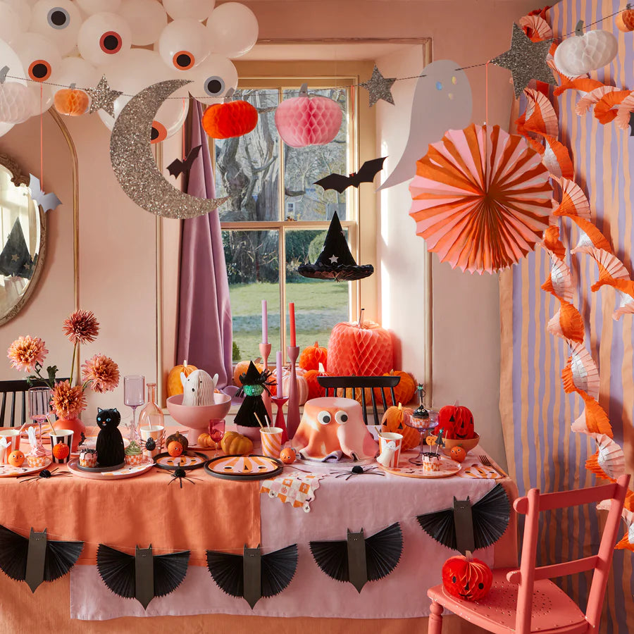 A festive Halloween-themed table setup featuring a mix of orange, black, and pink decorations. The table is adorned with pumpkins, candles, a black cat figurine, and ghost-shaped hanging ornaments. A crescent moon and stars hang above the table along with a spooky Meri Meri Bat Garland (Final Sale), with a window in the background.